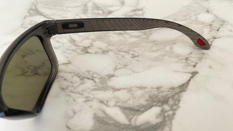 May include: Dark gray sunglasses with a rectangular frame and tinted lenses. The temples are a lighter gray with a textured design and a small red accent at the end. The sunglasses are set against a marble background.