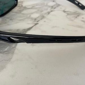 May include: Black sunglasses with a sleek, modern design. The frames are glossy and curved, with a subtle cutout detail on the temples. The lenses have a reflective, blue-green tint, suggesting polarized protection.