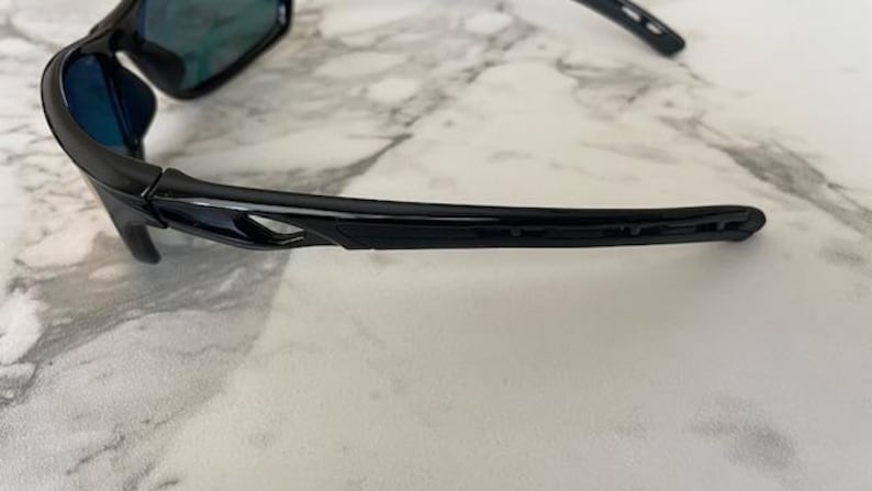 May include: Black sunglasses with a sleek, modern design. The frames are glossy and curved, with a subtle cutout detail on the temples. The lenses have a reflective, blue-green tint, suggesting polarized protection.