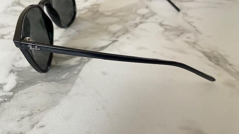Puede incluir: Gafas de sol negras con lentes oscuras. El logotipo de Ray-Ban es visible en la patilla. Las gafas de sol est&aacute;n sobre un fondo de m&aacute;rmol.