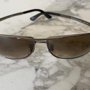 May include: Sunglasses with a dark metal frame and gradient brown lenses. The sunglasses have a sleek, modern design with a curved shape. The Ray-Ban logo is visible on the left lens. The arms are black.