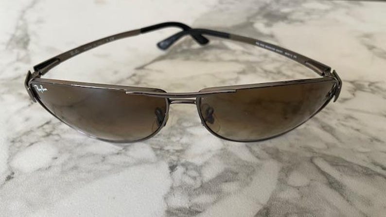 May include: Sunglasses with a dark metal frame and gradient brown lenses. The sunglasses have a sleek, modern design with a curved shape. The Ray-Ban logo is visible on the left lens. The arms are black.