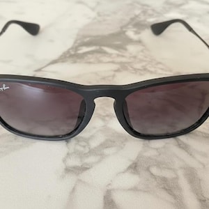 Puede incluir: Gafas de sol negras con acabado mate. Las lentes tienen un degradado de oscuro a claro. El logotipo de Ray-Ban es visible en la lente izquierda. La montura tiene una forma rectangular cl&aacute;sica.