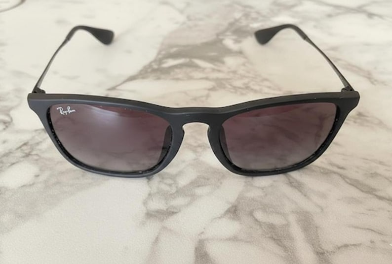 Puede incluir: Gafas de sol negras con acabado mate. Las lentes tienen un degradado de oscuro a claro. El logotipo de Ray-Ban es visible en la lente izquierda. La montura tiene una forma rectangular cl&aacute;sica.