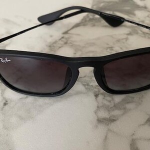 Puede incluir: Gafas de sol negras con lentes oscuras. Las gafas tienen una montura negra mate y patillas negras finas. El logotipo de Ray-Ban es visible en la lente izquierda. Las gafas descansan sobre una superficie clara.