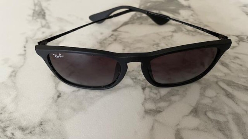 Puede incluir: Gafas de sol negras con lentes oscuras. Las gafas tienen una montura negra mate y patillas negras finas. El logotipo de Ray-Ban es visible en la lente izquierda. Las gafas descansan sobre una superficie clara.