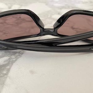 May include: Black sunglasses with a glossy finish, featuring a square frame and tinted lenses. The arms of the sunglasses are curved and have a subtle logo detail. The sunglasses are resting on a light-colored surface.