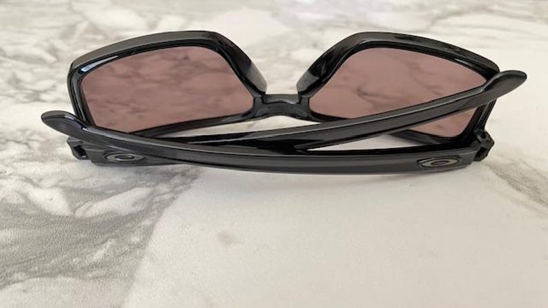 May include: Black sunglasses with a glossy finish, featuring a square frame and tinted lenses. The arms of the sunglasses are curved and have a subtle logo detail. The sunglasses are resting on a light-colored surface.