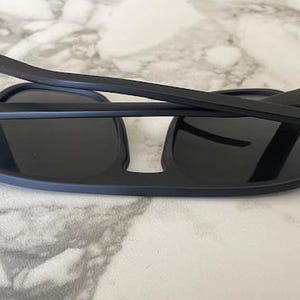 Puede incluir: Gafas de sol negro mate con lentes oscuras y montura plegable. Las patillas presentan dos finas rayas horizontales grises cerca de las bisagras, unidas a la estructura principal mediante herrajes met&aacute;licos de color plateado.