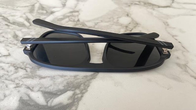 Puede incluir: Gafas de sol negro mate con lentes oscuras y montura plegable. Las patillas presentan dos finas rayas horizontales grises cerca de las bisagras, unidas a la estructura principal mediante herrajes met&aacute;licos de color plateado.