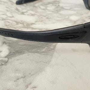 Może przedstawiać: Czarne okulary przeciwsłoneczne Oakley z zielonymi szkłami. Oprawka jest matowa czarna z logo Oakley widocznym na zauszniku. Okulary przeciwsłoneczne leżą na jasnej, marmuropodobnej powierzchni.