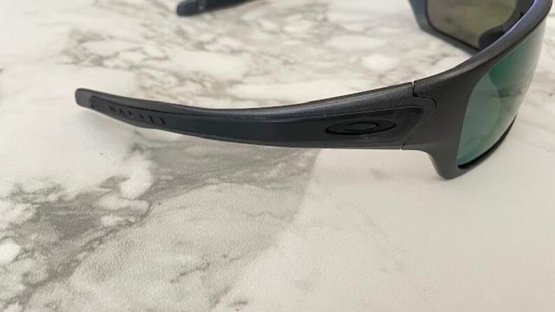 Może przedstawiać: Czarne okulary przeciwsłoneczne Oakley z zielonymi szkłami. Oprawka jest matowa czarna z logo Oakley widocznym na zauszniku. Okulary przeciwsłoneczne leżą na jasnej, marmuropodobnej powierzchni.