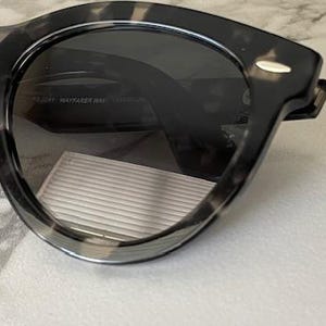 Puede incluir: Primer plano de gafas de sol con montura moteada gris oscuro y negra. Las lentes son de color gris oscuro y las gafas de sol tienen una forma cl&aacute;sica y redondeada. La montura tiene un patr&oacute;n sutil y texturizado.