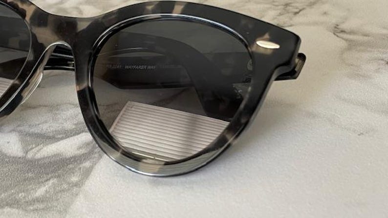 Puede incluir: Primer plano de gafas de sol con montura moteada gris oscuro y negra. Las lentes son de color gris oscuro y las gafas de sol tienen una forma cl&aacute;sica y redondeada. La montura tiene un patr&oacute;n sutil y texturizado.