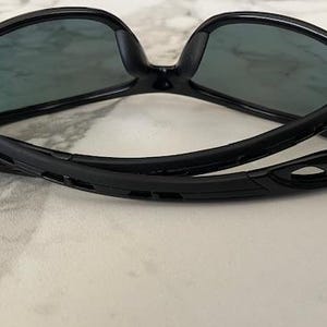 May include: Black sunglasses with dark lenses. The sunglasses have a sleek, modern design with a curved frame and temples. The lenses are a dark gray color, and the frame is glossy black.