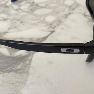 May include: Black sunglasses with a matte finish. The frame features a silver logo on the temple and a hinge with silver rivets. The lenses are dark, and the overall design is sleek and modern.