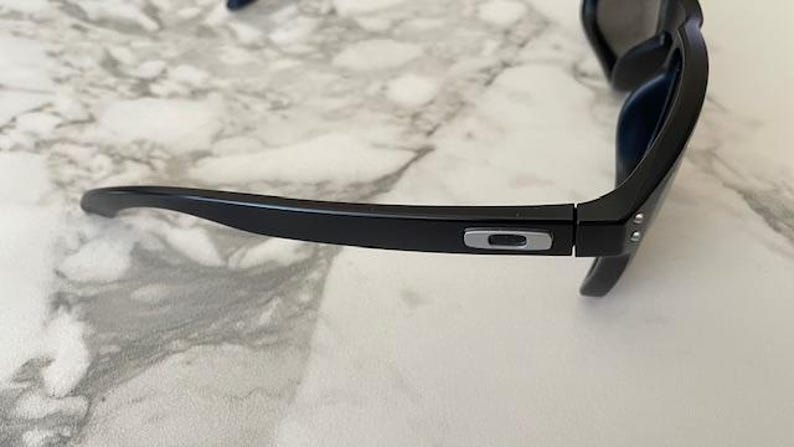 May include: Black sunglasses with a matte finish. The frame features a silver logo on the temple and a hinge with silver rivets. The lenses are dark, and the overall design is sleek and modern.