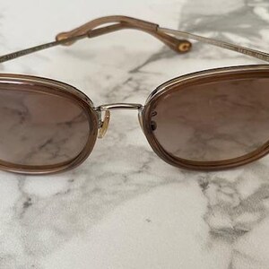 May include: Brown cat-eye sunglasses with a metal bridge and temples. The lenses are a gradient brown color. The frame is a light brown color. The sunglasses are on a white and gray marble surface.
