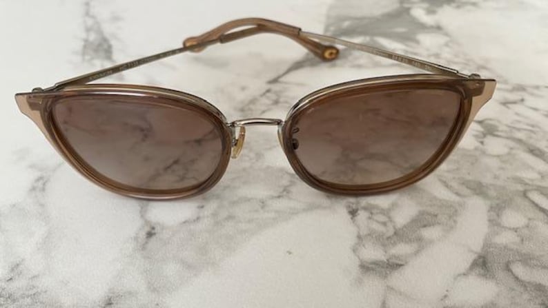May include: Brown cat-eye sunglasses with a metal bridge and temples. The lenses are a gradient brown color. The frame is a light brown color. The sunglasses are on a white and gray marble surface.