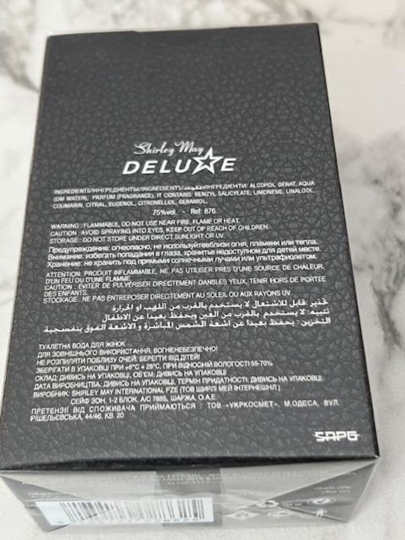 May include: Black box of Shirley May Deluxe perfume. The box has a textured surface and features the brand name in gold lettering. The ingredients and warnings are printed on the box.