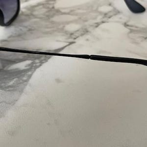 May include: Black sunglasses with dark lenses. The frame is black with a thin, black arm extending to the right. The Ray-Ban logo is visible on the arm. The sunglasses are resting on a white and gray marble surface.