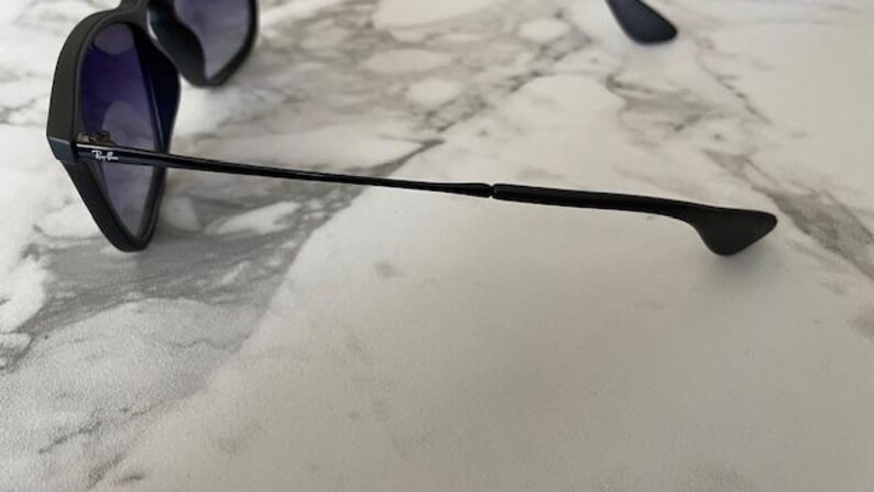 May include: Black sunglasses with dark lenses. The frame is black with a thin, black arm extending to the right. The Ray-Ban logo is visible on the arm. The sunglasses are resting on a white and gray marble surface.