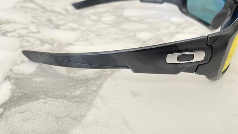 Puede incluir: Gafas de sol negras con un logotipo plateado en la patilla. Las lentes tienen un tinte amarillo y las monturas son negras. Las gafas de sol est&aacute;n dise&ntilde;adas para deportes o actividades al aire libre.