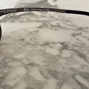 May include: Close-up of Ray-Ban sunglasses. The sunglasses have a dark metal frame and brown tinted lenses. The temple arm is black and has the text "MADE IN ITALY CE". The Ray-Ban logo is visible on the lens.