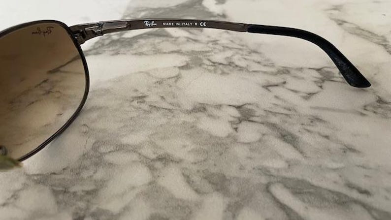 May include: Close-up of Ray-Ban sunglasses. The sunglasses have a dark metal frame and brown tinted lenses. The temple arm is black and has the text "MADE IN ITALY CE". The Ray-Ban logo is visible on the lens.