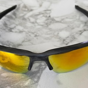 Puede incluir: Gafas de sol negras con lentes doradas. Las gafas tienen un dise&ntilde;o elegante y moderno con montura negra y patillas curvas. Las lentes son de color dorado vibrante, reflejando la luz.
