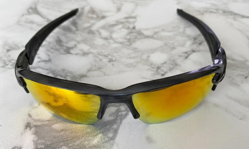 Puede incluir: Gafas de sol negras con lentes doradas. Las gafas tienen un dise&ntilde;o elegante y moderno con montura negra y patillas curvas. Las lentes son de color dorado vibrante, reflejando la luz.