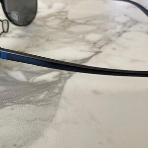 May include: Black aviator sunglasses with dark lenses and a sleek, modern design. The frames are black with a blue accent on the temples. The Oakley logo is visible on the temple.