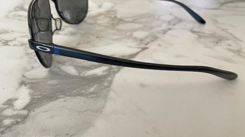 May include: Black aviator sunglasses with dark lenses and a sleek, modern design. The frames are black with a blue accent on the temples. The Oakley logo is visible on the temple.