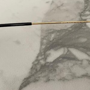 May include: Close-up of sunglasses with gold-colored frames and dark lenses. The temple arm is black at the end, transitioning to gold. The glasses are set against a light-colored, marbled surface.