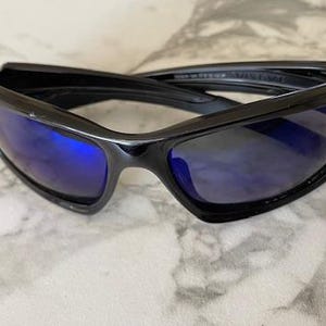 May include: Black sunglasses with blue lenses. The sunglasses have a sleek, modern design with a glossy finish. The lenses are a vibrant blue color, and the frames are a solid black. The sunglasses are likely for outdoor wear.