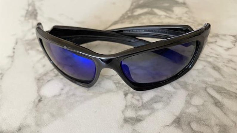 May include: Black sunglasses with blue lenses. The sunglasses have a sleek, modern design with a glossy finish. The lenses are a vibrant blue color, and the frames are a solid black. The sunglasses are likely for outdoor wear.
