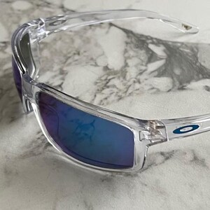 May include: Clear-framed sunglasses with blue-tinted lenses. The sunglasses have a sleek, modern design with a curved frame and a small logo on the temple. The lenses reflect a gradient of blue and purple hues.
