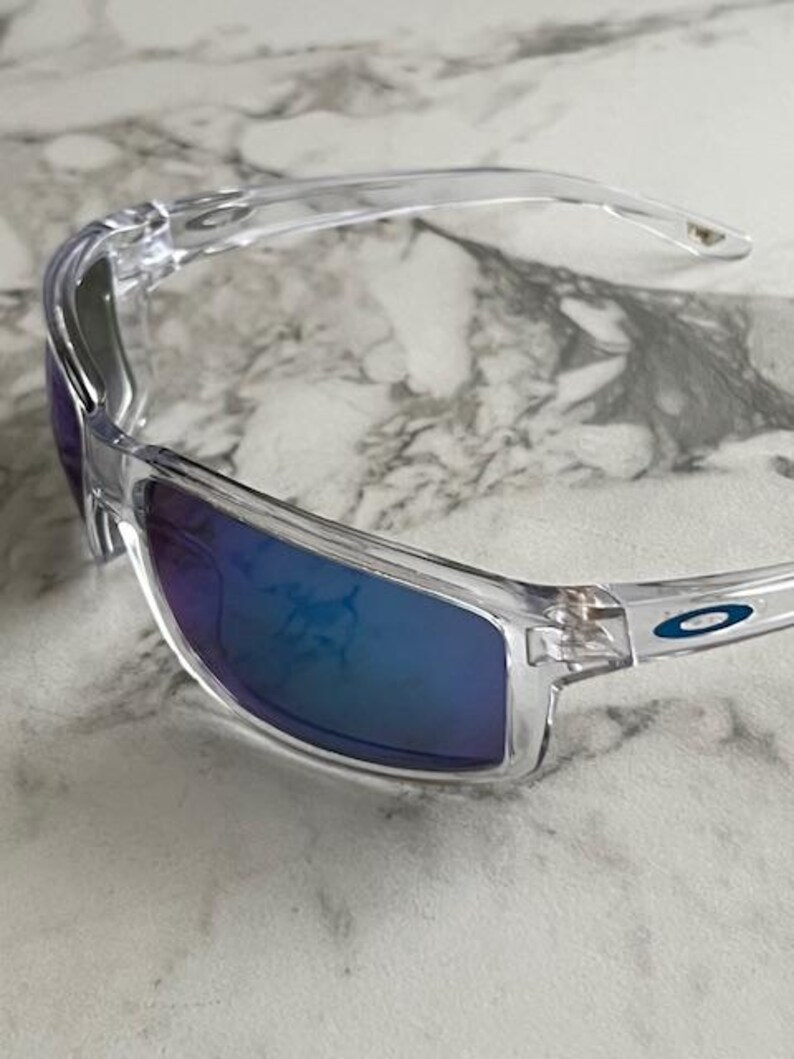 May include: Clear-framed sunglasses with blue-tinted lenses. The sunglasses have a sleek, modern design with a curved frame and a small logo on the temple. The lenses reflect a gradient of blue and purple hues.