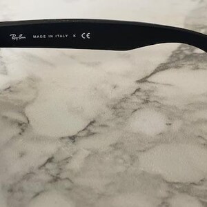 May include: Black sunglasses with a dark green lens. The temple arm features the Ray-Ban logo, the text "MADE IN ITALY", and the CE mark. The sunglasses are set against a marble background.
