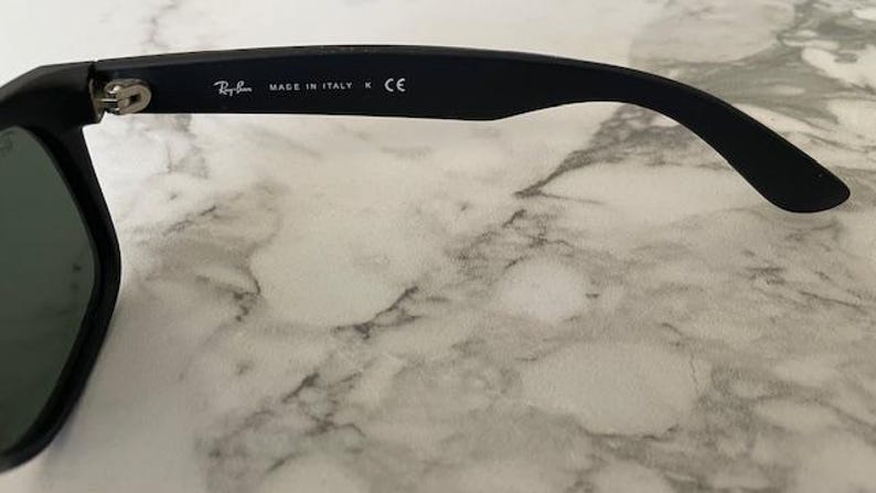 May include: Black sunglasses with a dark green lens. The temple arm features the Ray-Ban logo, the text "MADE IN ITALY", and the CE mark. The sunglasses are set against a marble background.
