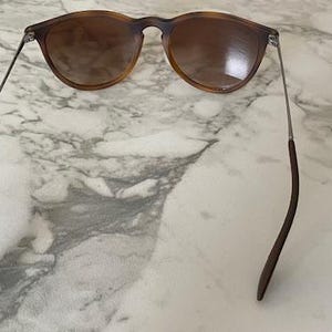 May include: Sunglasses with a brown tortoiseshell frame and tinted lenses. The sunglasses have thin, silver-colored arms that connect to the curved, dark brown temples. The glasses are resting on a white and gray marble surface.