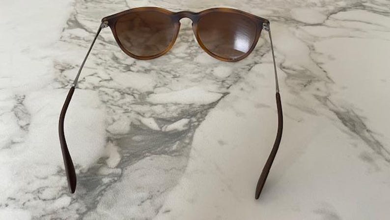 May include: Sunglasses with a brown tortoiseshell frame and tinted lenses. The sunglasses have thin, silver-colored arms that connect to the curved, dark brown temples. The glasses are resting on a white and gray marble surface.