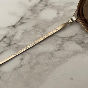 May include: Close-up of a pair of sunglasses with a light gold and brown frame. The temple arm is inscribed with "565413 (Light Gold/Brown) 54▢20 140 2N". The lenses are a gradient brown color. The background is a marble pattern.