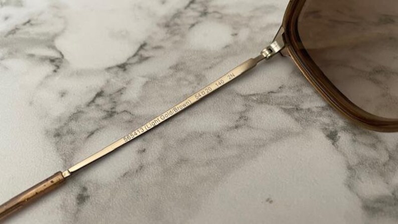 May include: Close-up of a pair of sunglasses with a light gold and brown frame. The temple arm is inscribed with "565413 (Light Gold/Brown) 54▢20 140 2N". The lenses are a gradient brown color. The background is a marble pattern.