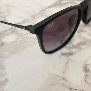 Puede incluir: Gafas de sol negras con lentes degradadas y el logotipo de Ray-Ban en la montura. Las gafas de sol est&aacute;n sobre una superficie de m&aacute;rmol blanco y gris.