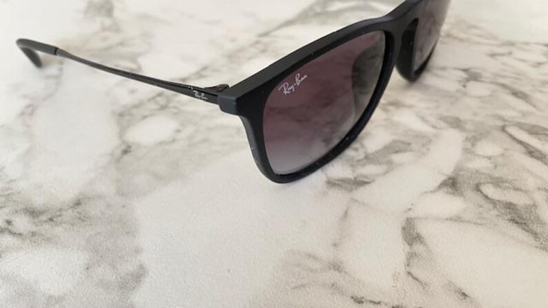Puede incluir: Gafas de sol negras con lentes degradadas y el logotipo de Ray-Ban en la montura. Las gafas de sol est&aacute;n sobre una superficie de m&aacute;rmol blanco y gris.