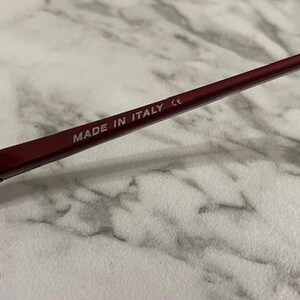 May include: Close-up of burgundy sunglasses with dark lenses. The frame is a deep red color, and the text "MADE IN ITALY" is printed on the temple arm. The sunglasses are set against a light-colored background.