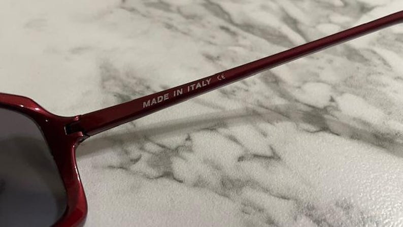 May include: Close-up of burgundy sunglasses with dark lenses. The frame is a deep red color, and the text "MADE IN ITALY" is printed on the temple arm. The sunglasses are set against a light-colored background.