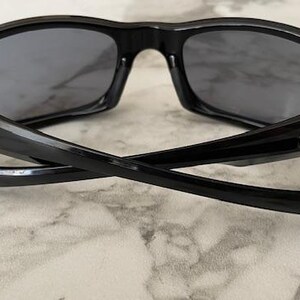 K&ouml;nnte beinhalten: Schwarze Sonnenbrille mit dunkelgrauen Gl&auml;sern. Die Sonnenbrille hat ein schlankes, modernes Design mit einem geschwungenen Rahmen und B&uuml;geln. Die B&uuml;gel sind gekreuzt und zeigen die Innendetails der Scharniere.