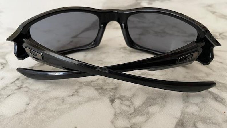 K&ouml;nnte beinhalten: Schwarze Sonnenbrille mit dunkelgrauen Gl&auml;sern. Die Sonnenbrille hat ein schlankes, modernes Design mit einem geschwungenen Rahmen und B&uuml;geln. Die B&uuml;gel sind gekreuzt und zeigen die Innendetails der Scharniere.